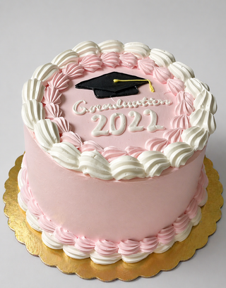 The Cap & Boarders cake features pink frosting, white icing borders, a black graduation cap decoration on top, and “Graduation 2021” written in white icing, all presented on a gold cake board.