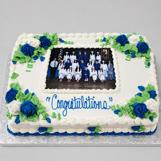 A rectangular cake decorated with blue, white, and green icing flowers, topped with a "Photo Sheet" featuring a group graduation photo and "Congratulations" written in blue icing.