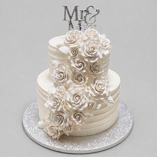 Wedding Cascade: A two-tiered white bridal cake adorned with cascading fondant roses and leaves, topped with a silver "Mr & Mrs" sign, presented on a silver-patterned board—perfect for your special day.