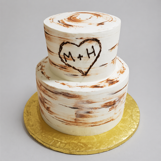 Heart Engrave is a two-tiered white cake with rustic brown brush strokes, perfect for bridal or engagement celebrations. Featuring a heart engrave design and M + H initials, it sits elegantly on a gold board.