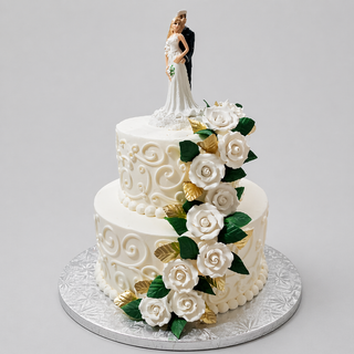 The Newlywed Tier is a two-tier white bridal cake with intricate swirl icing, cascading white fondant roses, green and gold leaves, and a bride and groom figurine on top, all elegantly presented on a silver board.
