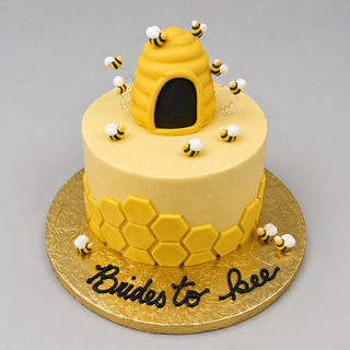 The "Bees Flying" cake features yellow icing, hexagon shapes, fondant bees, a Fondant Hive beehive topper, and “Brides to bee” in black icing on the base, all displayed on a gold board.