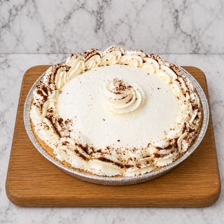 A Chocolate Cream Pie with whipped cream, chocolate shavings, and decorative swirls sits on a wooden board against a marble background.