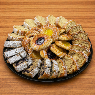 The Breakfast Loaf & Danish Platter features sliced banana bread, blueberry loaf with powdered sugar, and mini iced danishes, all arranged on a round black tray atop a wooden table.