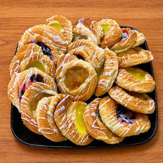 A Mini Danish Platter with lemon, blueberry, and apple fruit danishes topped with white icing is displayed on a black plate set on a wooden surface.