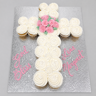 The Cupcake Religious cake features cupcakes arranged in a cross shape with white frosting, pink rose decorations, and "God Bless Lexa Margaret" written in pink on a silver board—ideal for religious celebrations.