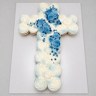 The Blessed Cupcakes cake is cross-shaped with white swirled frosting, light blue accents, and blue and white icing roses, all displayed on a white rectangular board.