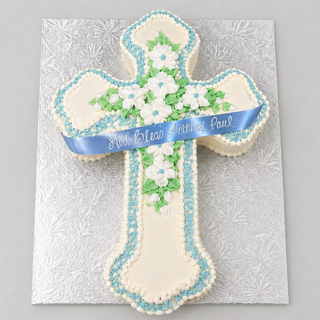 The Swirled Cross cake features a cross shape with white flowers, green leaves, blue and white icing borders in a Fancy Scroll Cut design, topped with a blue "God Bless Anthony Paul" ribbon on a silver-patterned board.
