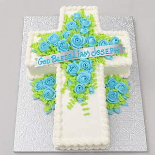 The Religious Cutout cake is cross-shaped, decorated with blue roses, green leaves, and a banner that reads "God Bless Liam Joseph," elegantly displayed on a textured silver board.