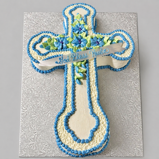 A Fancy Cross cake decorated with blue and green flowers, blue piping, and a white "God Bless Tanto" banner sits on a textured silver board, perfect for a special religious celebration.
