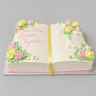 The Religious Book cake is shaped like a book and decorated with pink and yellow icing flowers, green leaves, and a yellow ribbon. "God Bless Isabella Elizabeth" is written in pink icing on the left side, and the cake sits on a silver tray.