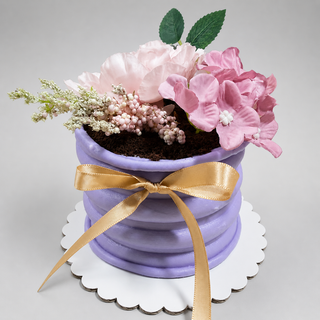 The 4” Vase Cake resembles a purple flower pot, topped with realistic pink and white edible flowers, cocoa crumb “soil,” and a gold ribbon, all presented on a white scalloped cake board.
