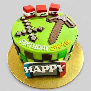 The Minecraft Essentials cake is round and decorated with TNT blocks, a pickaxe, green blocks, "Birthday Samir" on top, a HAPPY sign on the side, and sits on a gold board.