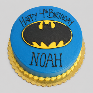 A blue Batman Logo kids superhero birthday cake with a black and yellow Batman emblem, decorated with "Happy 4th Birthday Noah" in black icing on a gold scalloped board.