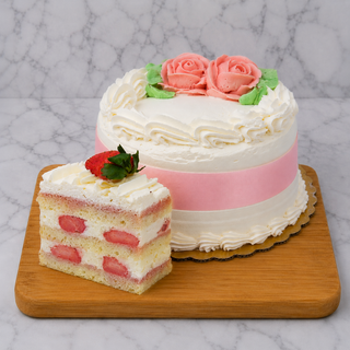 Strawberry Shortcake: A round layered cake with white frosting, pink ribbon, and rose decorations sits on a wooden board. In front, a slice of sponge cake filled with strawberries and homemade whip cream is topped with a fresh strawberry.