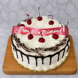 The Black Forest cake is a round chocolate creation with white frosting, chocolate ganache drips, whipped cream swirls, cherries on top, and a pink ribbon reading "Happy 65th Birthday!" in white icing. It’s presented on a wooden board.