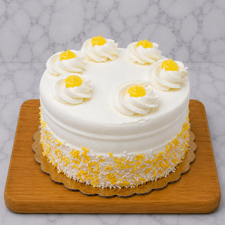 The Lemon Layer Cake features a round shape with white buttercream frosting, topped with six swirls of white and yellow cream and decorated with yellow and white sprinkles around the base. It’s presented on a wooden board with a marble background.