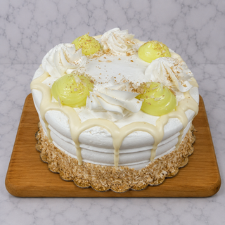 The Key Lime Layer Cake—with white frosting, yellow cream dollops, white chocolate drips, crumbs, and swirls—rests on a wooden board against a marble background.