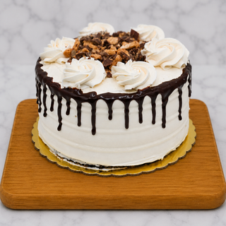 The Peanut Butter Layer Cake features white frosting, chocolate ganache drips, whipped cream swirls, and chopped chocolate pieces on top. Presented on a wooden board against a marble background.