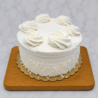 The White Layer Cake is a round cake with white buttercream, six large swirls on top, white sprinkles on the sides, set on a gold cake base atop a wooden board against a light marble background.