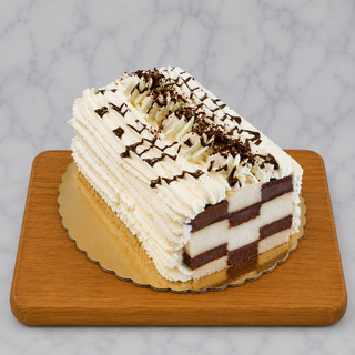 The Checkerboard Cake features chocolate and vanilla stripes, topped with whipped cream and rich chocolate ganache, elegantly displayed on a gold cake board atop a wooden tray with a marble background.