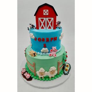 The Barn Tier is a two-tiered farm-themed birthday cake with a red barn topper, fondant animals, white fencing, and "Joseph 1" in red letters on the blue upper tier. The green lower tier features more animals and flowers.