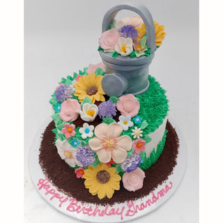The Garden Watering cake is a round cake featuring colorful fondant flowers, green icing grass, a gray watering can topper, chocolate crumb edges, and "Happy Birthday Grandma" written in pink icing.