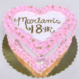 The Heart Birthday cake features a heart shape, pink frosting, gold decorative balls, and "Mariam's 18th" in gold icing on top, all presented on a gold cake board.