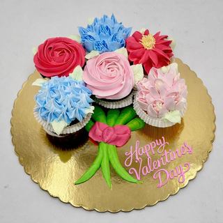 The Valentines Bouquet features six cupcakes decorated as colorful flowers, arranged like a bouquet on a gold board with "Happy Valentines Day" in pink icing and green icing stems and ribbon.