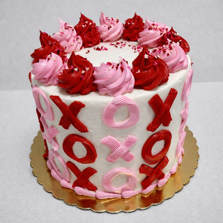 The Valentines XO cake is round, decorated with pink and red “X” and “O” icing on the sides, topped with swirls of pink and red frosting, and displayed on a gold cake board against a plain background.