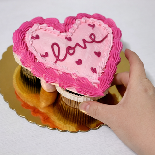 A hand holds the Valentines Pull-Apart, a heart-shaped cake with pink icing, smaller hearts, and “love” in cursive. The cake rests on a gold tray against a white background.