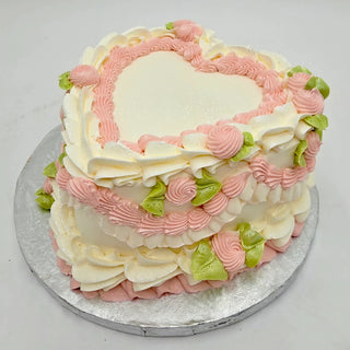 The Valentines Vintage is a heart-shaped cake with white, pink, and green frosting, piped roses and leaves, displayed on a silver cake board against a white background.