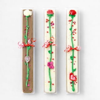 Three rectangular Valentines Rose Cakes in clear packaging, each adorned with an icing long-stemmed rose and ribbons, plus heart and flower accents, all set against a white background.