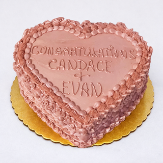 The Heart Engage cake is a heart-shaped pink engagement cake with decorative frosting on a gold board, featuring “CONGRATULATIONS CANDACE + EVAN” written in handwritten-style icing on top.