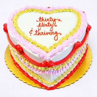 The Colorful Heart is a specialty birthday cake with pastel pink, yellow, and red frosting borders and "thirty, flirty & thriving" written in red icing on top, finished on a gold cake board.