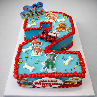 The Paw Patrol Number cake is shaped like a 5 and decorated with Paw Patrol characters, toy bones, a small fire truck, red icing borders, and a “Happy Birthday” sign on a white board.