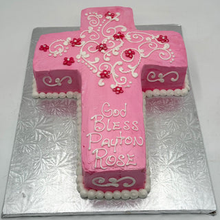 The Pink Cross cake is shaped like a cross and decorated with white icing designs and small red flowers, featuring "God Bless Payton Rose" on top. It is presented on a silver patterned board.