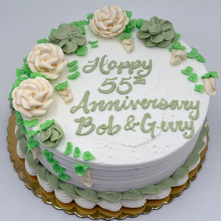 Pure Elegance is a round white cake with green and white icing flowers, leaves, and trim. "Happy 55th Anniversary Bob & Gary" is written in green icing. The cake sits on a gold-colored board.