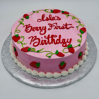 A round pink cake called Berry Cute, with "Lolas Berry First Birthday" in red icing, decorated with piped strawberries and green vines on a silver cake board, finished with white frosting along the base.
